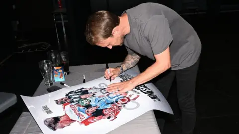 Doncaster Rovers A man in grey, with one arm tattooed heavily, bends down at a table to sign a movie poster.
