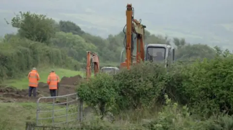 BBC POOL A screenshot of television footage of the ICLVR search for Captain Robert Nairac. Two men dressed in high visibility orange jackets stand in a field next to an open gate. Two mechanical backhoes are also on the ground and much of the grass has been dug up and turned over for examination.