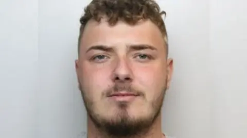 A police custody image of a man with short brown hair and a brown close-trimmed beard.