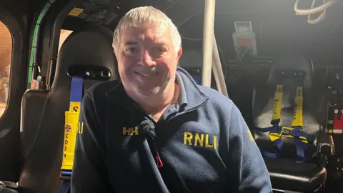 Dave Whiteley, a man with white hair wearing a navy fleece with RNLI written on it in yellow. He is in a lifeboat and there are seats behind him.