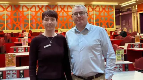 A woman with short dark brown hair and wearing a black jumper is standing alongside a man with short hair and glasses who is wearing a light blue shirt and beige trousers. The are sanding in a bingo hall with red seating and white tables. A man is sat at a table in the background.