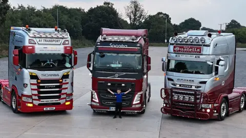 Family Handout A boy in a royal blue t-shirt and dark trousers standing in front of the middle one of three trucks, with his arms spread wide. The red, maroon and silver trucks, which are not carrying loads, are standing on a wide forecourt with trees in the background.