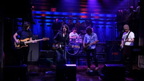 Getty Images John on stage with the band during a live performance. He is wearing a brown beanie hat and white t-shirt.