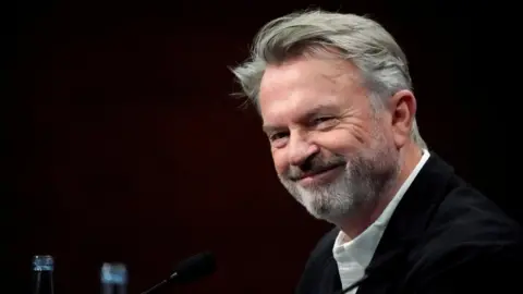Sam Neill smiles against a black background