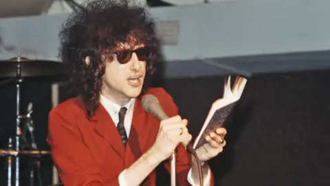 Getty Images John Cooper Clarke in an orange suit talking in an animated way into a microphone, holding up a book with one hand, circa 1990