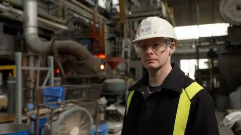 Image of scientist Dr Cyrille Dunant standing in front of an electric arc furnace at the Middlesborough Materials Processing Laboratory.  The furnace is red hot.