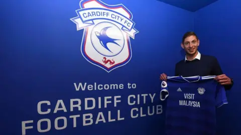 Getty Images A young man with brown hair, wearing a white shirt and dark jumper. He is holding a blue Cardiff City football shirt and is stood in front of a blue, Cardiff City branded wall which reads "Welcome to Cardiff City Football Club". He is smiling and looking at the camera.