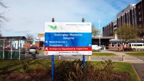 A blue-and-white sign outside Darlington Memorial Hospital. The sign points to the main entrance and emergency department in red. In the background, to the right, is a large, brick building.