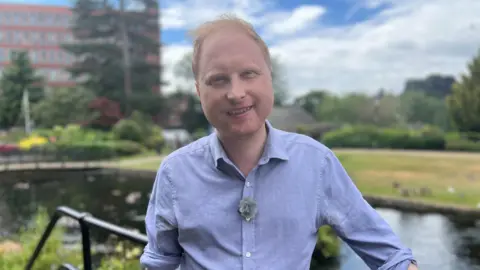 Man with short red hair and blue shirt stood on a bridge infront of river in park area