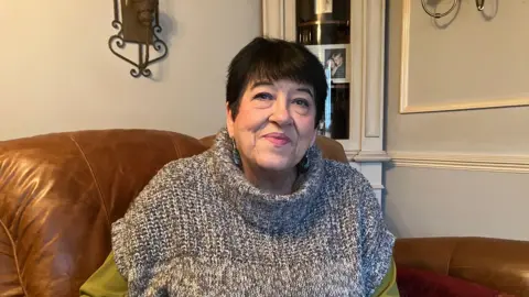 Amy Holmes/BBC A woman sits on a brown leather sofa in a living room, wearing a grey knitted sleeveless sweater over a green long‑sleeve top, with a cabinet and wall decor in the background.