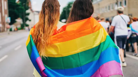 Two women with the rainbow flag wrapped around them, standing on a road behind other people as a parade takes place.