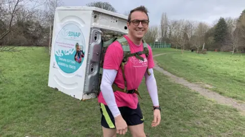 A man dressed in running gear - including a bright pink T-shirt with a long sleeved white top underneath and black shorts with a neon green stripe - smiles as he stands in a park with a white washing machine strapped to his back. There is a poster on the side of the washing machine asking people to donate to the charity he is raising money for. He is wearing glasses.