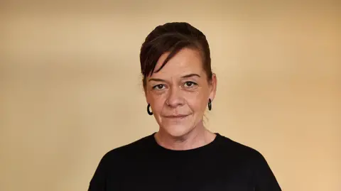 Shelter Amanda Wall with her dark hair tied up, black earrings and a nose ring. She is wearing a black top and smiling. 