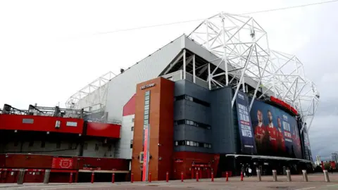 Getty Images The outside of Manchester United's Old Trafford. It is silver and red, with large white metal protruding from the top of it. There is a large black screen above its entrance with photos of players on it.