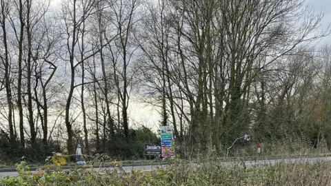 Side view of the site where the body was found. Police tape is caught in the trees either side of some advertising signage