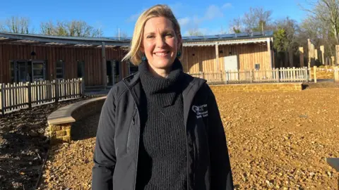 Rachel is stood in the middle of the shot, smiling at the camera. She's wearing a black jumper and black coat. Behind her is the wooden-clad building curving around the muddy stone circle.