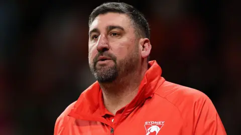 A man wears a red jacket emblazoned with a triangular "Sydney Swans" logo.