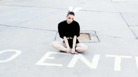DJ Boo is sat cross-legged next to a white 'No Entry' sign on the floor. She is wearing black shorts and a fluffy black top. On her head is a white headband with bunny ears on.
