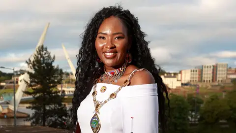 Shows Lilian Seenoi-Barr in a white dress wearing the mayoral chain and a colourful necklace and standing against the backdrop of the peace bridge.
