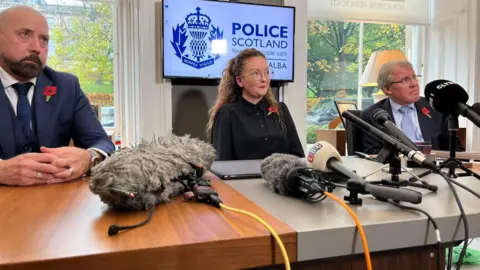 A bald man in a suit with a beard, a young woman with long curly hair and round glasses and a man with white hair and glasses sit left to right in a row behind a desk covered in microphones. There is a screen with Police Scotland behind them, between two windows looking on to greenery.