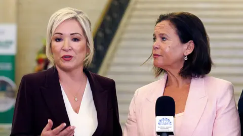 Michelle O'Neill,a woman with blonde hair tied back, and Mary Lou McDonald, a woman with short, dark hair, speak into microphones at Stormont
