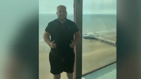 Richard Gallimore Richard standing in front of a window which shows a beach view. He is looking at the camera with his thumbs up and wears a black polo t-shirt with black shorts. 