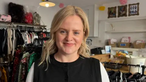 The picture shows a woman smiling at the camera. She has blonde, wavy shoulder length hair. She's wearing a white top which has been covered by a black waistcoat. She's standing in a shop. There are rails of clothes over her left shoulder, and shelves over her right shoulder. 