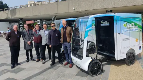 BCP Council Andrew Wood, catering manager at BCP Council, Joseph McMullen,Law Lecturer at Bournemouth University, Alain Simons, Senior Lecturer in Games Technology Programming at Bournemouth University, Richard Wintrip, Transport Policy Team Leader at BCP Council, Jamie, Front of House Host at BCP Council, and Ian Selby, Senior Travel Planning Officer at BCP Council. They are stood in front of and beside two of the brand new e-cargo vehicles.