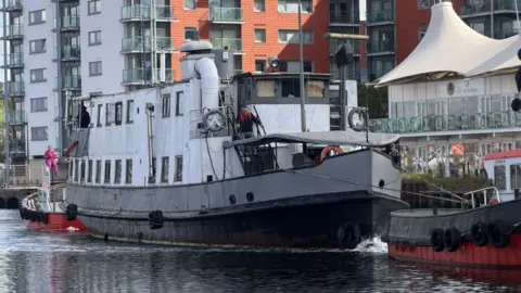 A white boat docked in a waterfront. A man can be seen walking down some steps on the front of the boat. Blocks of flats can be seen behind the boat.