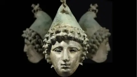 A photo of the Crosby Garrett Roman helmet; bronze sculpture of a face with curly hair and a pointy hat.