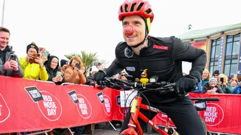 Radio 1 DJ Greg James is on his red bike on the starting line runway. Either side is a metal barrier lined with people waving. He is wearing all black cycling outfit and a red helmet and a red nose. 