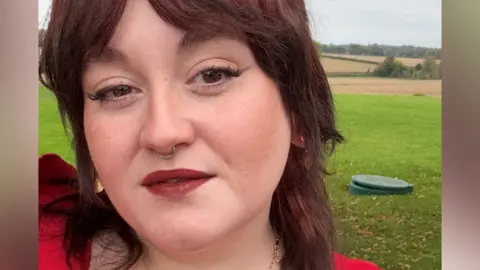 Frankie Bridger Frankie, a young woman with brown hair and brown eyes. She is wearing red-brown lipstick and light eye makeup. She is standing in a grassy area with fields in the background