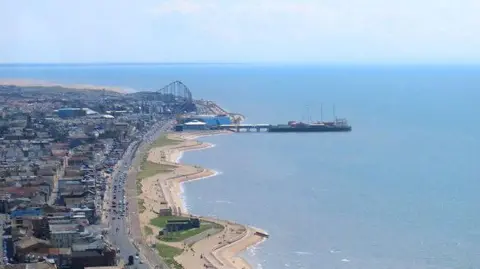 Geograph/Carroll Pierce Aerial view of South Pier jutting out on to the blue Irish Sea next to the Big One rollercoaster. The sandy beach lines the rest of the coast with the town on the left on a sunny day.