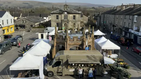 The town square in Kirkby Lonsdale is bordered by stone buildings, the one at the far end has tall arched windows and a clock above it. There is a pop-up market in the centre with white tents erected and a dark green food van. There is a stone structure in the centre with a stone cross atop it.