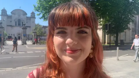 A girl with long died red hair and a fringe. She is wearing big gold earrings and a pink strappy top and is smiling while looking at the camera. In the background is an old university building and a main road with pedestrians walking past.