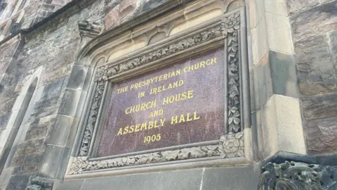 A sign built into a brick wall, reading: "The Presbyterian Church in Ireland, Church house and Assembly Hall 1905"
