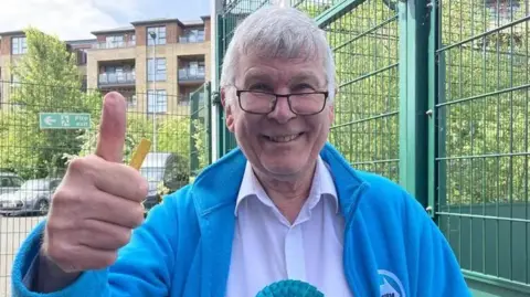 Mr Hopkins has grey hair and is wearing a blue jacket and white shirt with a blue rosette on. He is smiling and giving a thumbs up to the camera. He is stood next to a green metal fence and there is a block of flats behind him.