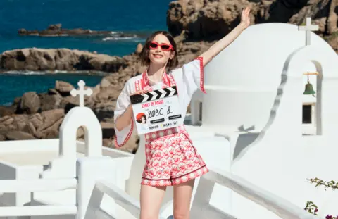 Lilly Collins stands with a clapperboard in a white t-shirt and red and white shorts. She extends one arm and wears red sunglasses. In the background is a white church and the sea of Greece.