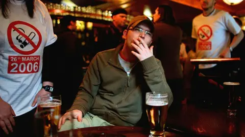 A man sitting at a table in a bar smokes a cigarette with a pint in front of him. Behind him, people are wearing t-shirts with a smoking ban symbol and the date of 26 March 2006