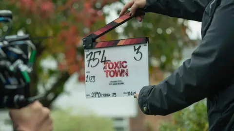 James Stack/Netflix A clapperboard with the words "Toxic Town" written in large red font is being held in front of a camera on set.