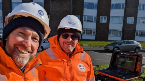 Two Southern Water team members smile for the camera next to a block of flats. They wear orange jackets and white hard hats. A high-tech laptop sits on top of equipment behind them.