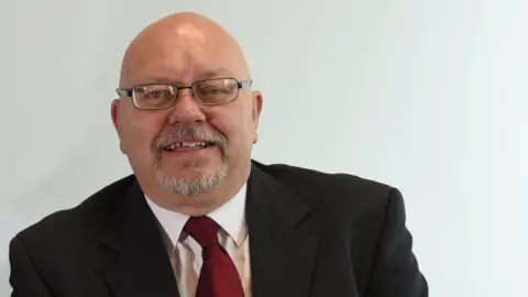 Glyn Jones looks at the camera. He is bald and has a greying goatee beard.  He is wearing rectangular shaped glasses.  He is wearing a black blazer, white shirt and burgundy coloured tie and is smiling.