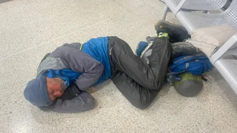 Nick Brooke Nick Brooke sleeping on a white and grey tiled floor next to a row of white metal seats in a ferry terminal. He is wearing blue and grey waterproof trousers and coat, hiking boots and a blue beanie hat. At his feet are his rucksack, sleeping bag and another white bag.