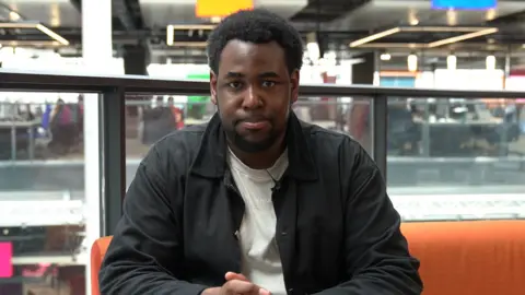 A man with a black jacker and a cream t shirt is looking directly into the camera. He is sat on an orange sofa, with glass barriers, desks and windows behind them. 
