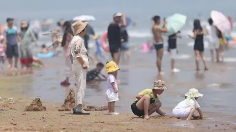 Getty Images A middle-aged woman wearing a brown hat standing on a beach. In front of her are three children playing with sand. In the background are many people on the crowded beach.