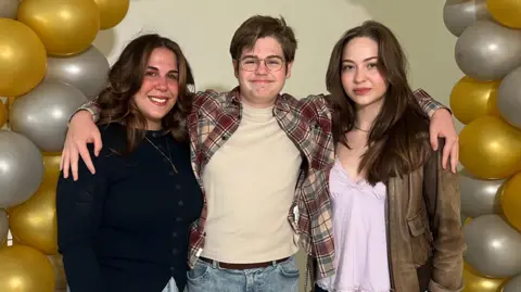 Mum Harriet stood with Sully and his sister Cordelia. They are all smiling at the camera and Sully has his arms wrapped around his mum and sister's shoulders.