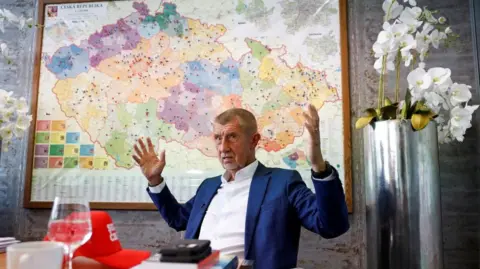 Reuters Andrej Babis gestures with both arms lifted, sitting in an office with a map of the Czech Republic behind him. There is a metallic-looking cylindrical vase of white flowers to the right of the picture. On the table in front of him are a red hat, a wine glass half full with clear liquid, and a white teacup. Babis is wearing a white shirt and navy blue jacket. He has grey stubble and short, cropped greying hair. 