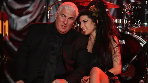 Getty Images Amy Winehouse with a black beehive hair style, and wearing a black dress, is sitting on the edge of the stage with her dad, he is wearing a black suit. He has grey hair. 