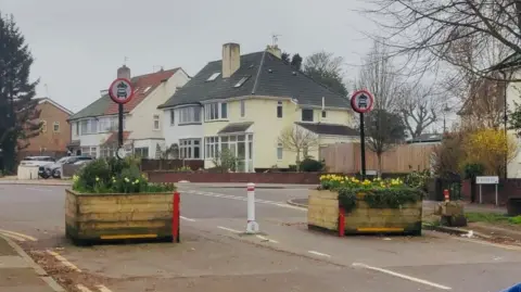 Image shows a road with two large wooden square planters filled with flowers either side of a white bollard.