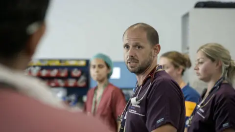 A male nurse captured in a still from the BBC series. He is looking at a colleague with a serious expression on his face, as several staff members stand around at a team briefing.